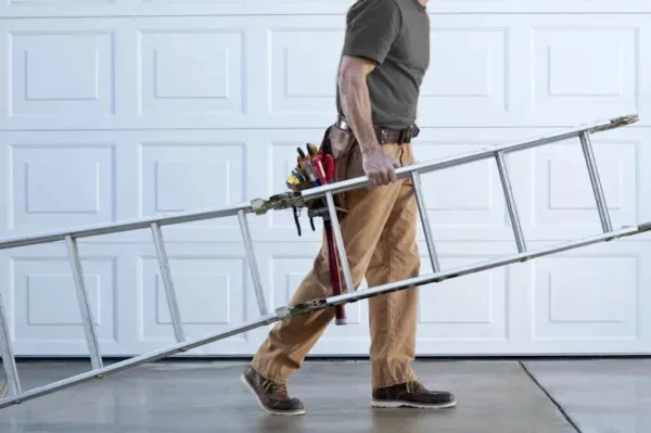 A professional technician wearing work clothes and a tool belt carries a long metal extension ladder, walking past a white sectional garage door with a raised panel design.