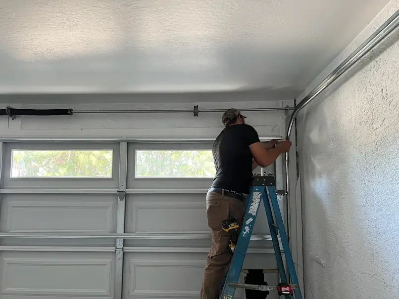 A bearded technician on a blue stepladder works on the curved top section of the metal garage door track rail inside a garage. The door is white and paneled with windows across the top section.