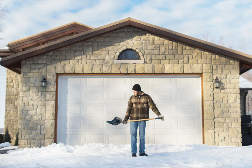 Man in plaid jacket and beanie holds a snow shovel in front of a large white garage door set into a house with beige stone siding during winter.